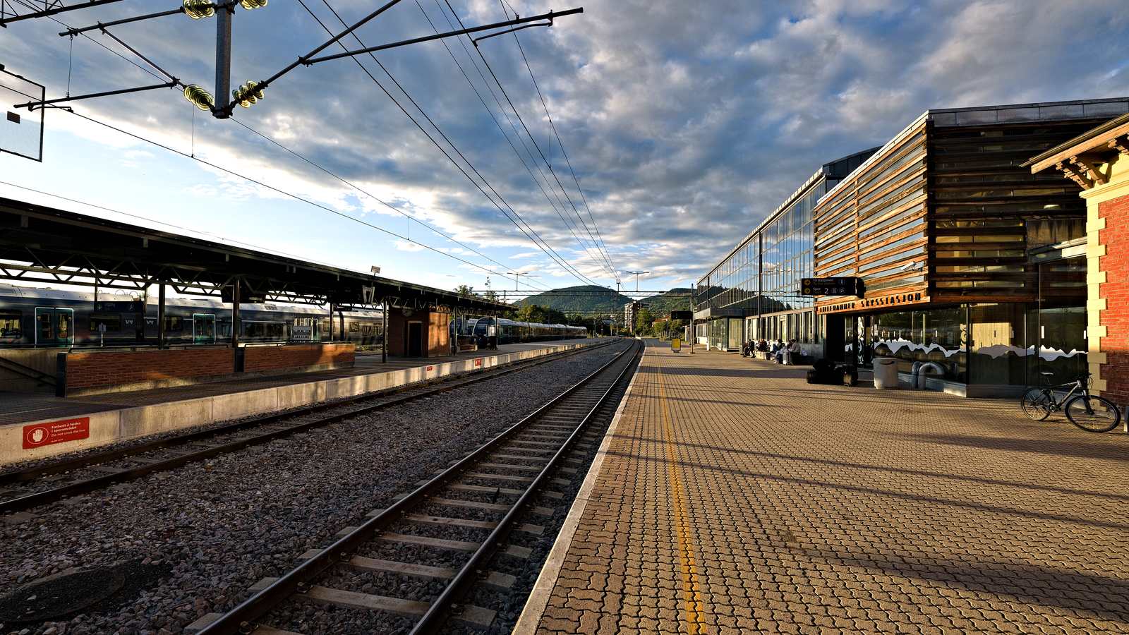 Plattformen på Lillehammer stasjon en sommerkveld, med tog ved perrongen og skysstasjonen til høyre